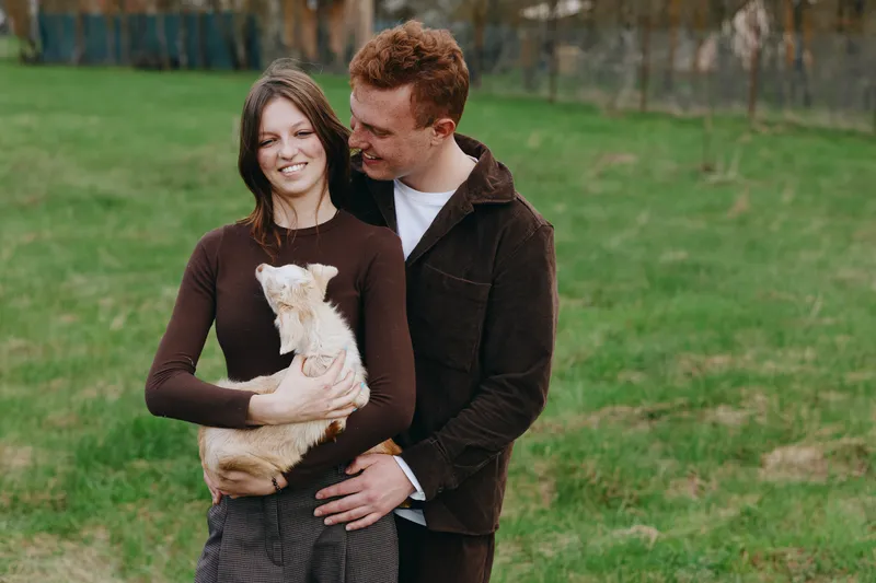 Cheerful couple outdoors, holding a goat kid, warm connection, casual attire, relaxed posture, sunlit meadow, soft breeze, natural colors, peaceful moment, candid affection