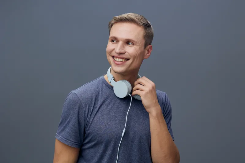 Smiling young man with short light brown hair wearing casual blue t-shirt holding white headphones against plain gray background studio portrait. People lifestyle concept, leisure, music, happiness, r