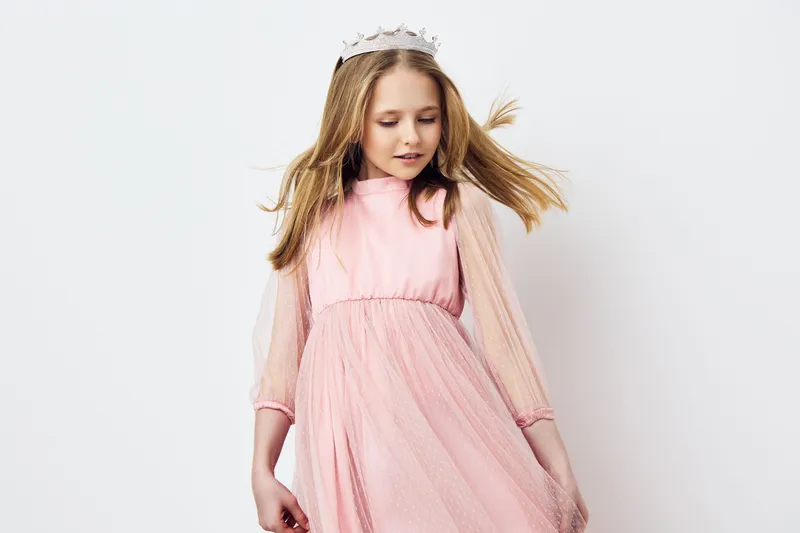 Young girl in a pink dress with long sleeves and a crown, smiling joyfully against a white background, embodying innocence and playfulness