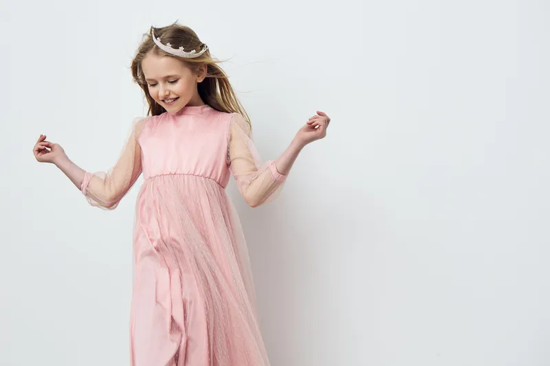 Delightful girl in a pink dress joyfully dancing with a tiara against a light background, showcasing childhood happiness and elegance