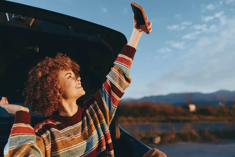 Curly woman lifestyle at sunset, holding a smartphone above an open car door, smiling outdoors with mountains and lake background, road trip mood, travel excitement summer air and freedom, today