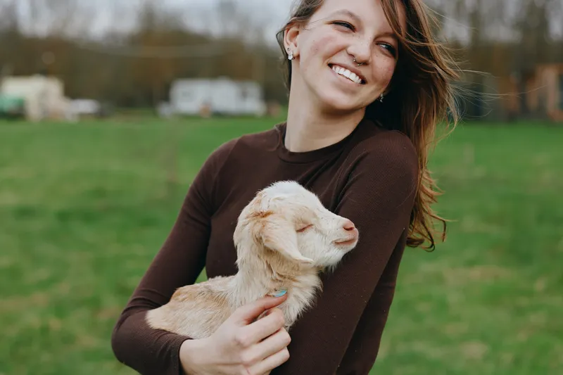 A cheerful woman outdoors, cradling a goat kid in a grassy field, captures a warm moment of countryside bonding. This lifestyle scene conveys happiness, gentle care, and connection with animals.