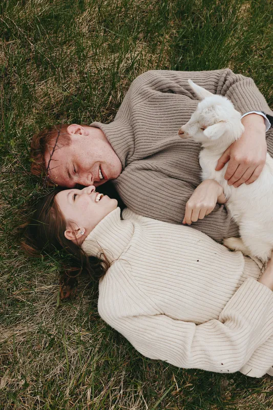Relaxed couple lying on the grass, sharing a gentle moment with a white goat kid. Soft natural light, cozy sweaters, outdoor scenery evokes wholesome lifestyle and bonding and shared joy.