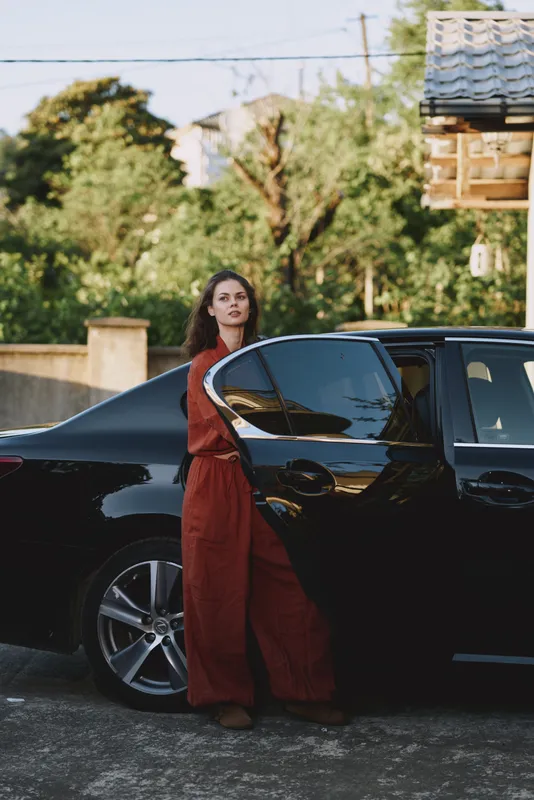 Woman lifestyle portrait beside a sleek black car in a sunlit driveway, wearing a red jumpsuit, looking at the camera with a relaxed confident vibe in a suburban setting and serene mood