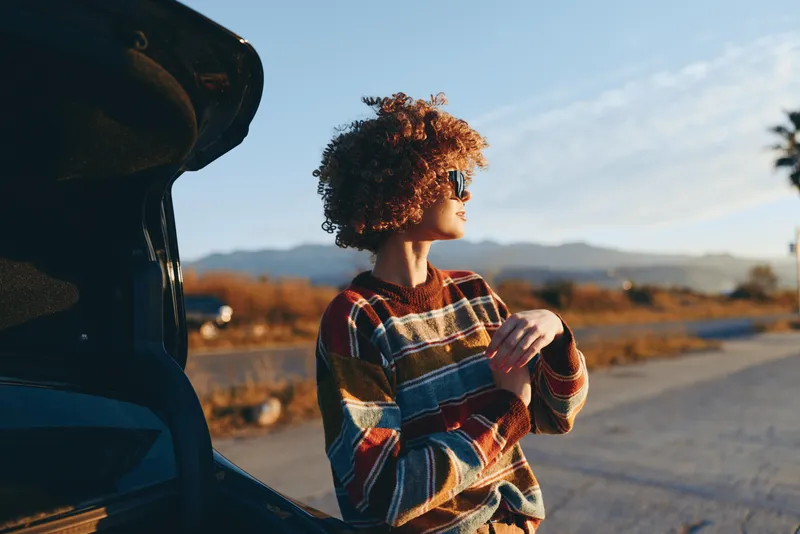 Woman outdoors lifestyle by car with curly hair, golden sunset light, plaid jacket, relaxed posture, standing near road, warm tones, distant mountains, peaceful mood, soft breeze, road ahead