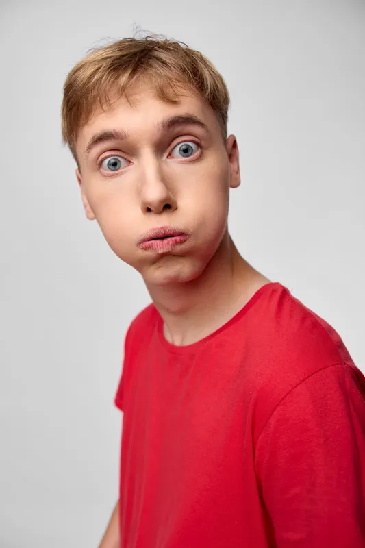 Surprised young man in a red shirt with wide eyes and puckered lips, studio setup, captured for humorous reaction and expressive stock imagery.