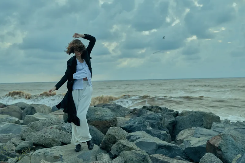 Fashion, woman, ocean, rocks, movement, wind, dress, jacket, coastline, dramatic leap along rough stones by the sea with moody clouds, outdoor styling for editorial use in fashion and lifestyle