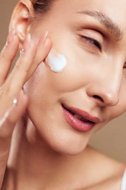 A close-up of a woman applying white cream on her cheek with fingers. The smooth skin and soft lighting highlight skincare and beauty care concepts.