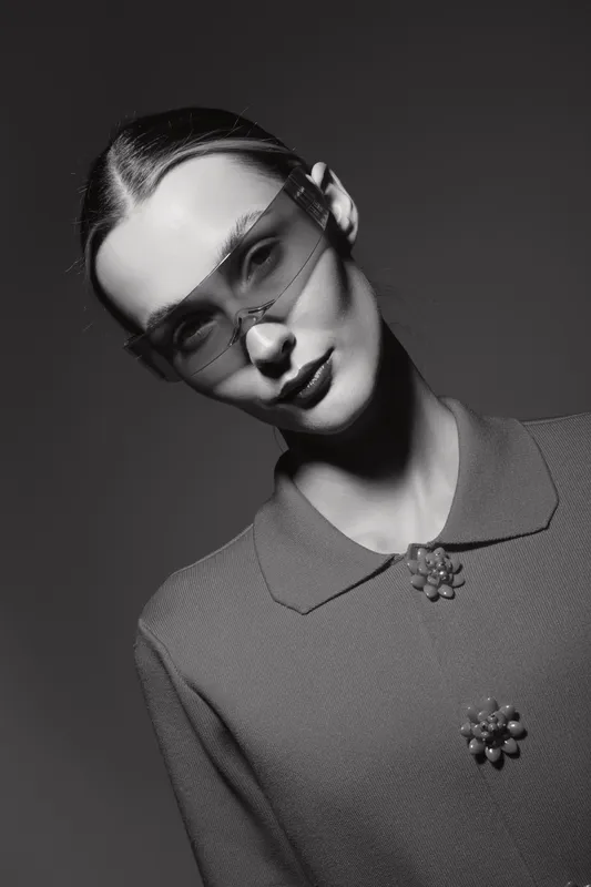 Portrait of a pale skinned young Caucasian woman wearing vintage glasses and textured coat with floral buttons in black and white studio lighting.