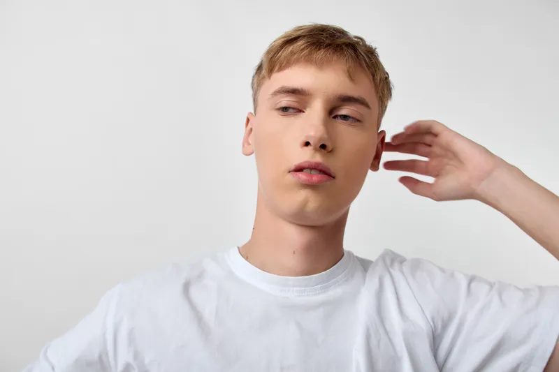 Young man wearing a white shirt in a minimal studio, with a pensive expression and a hand near the ear, creating a calm, thoughtful mood suitable for fashion and stock use