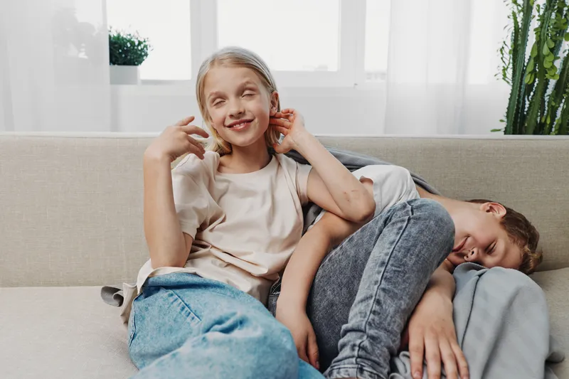 Two happy children relax on a sofa in a bright room, smiling and enjoying a cozy moment at home with natural light and casual clothing.