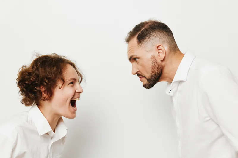 A business professional man stands against an isolated colored backdrop, displaying intense emotion as he faces a colleague in a tense, high energy moment of confrontation.