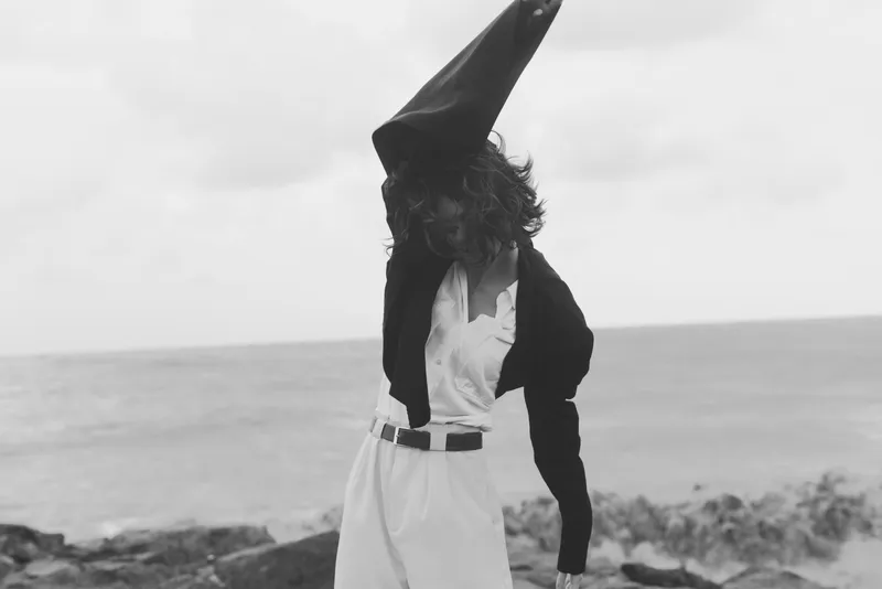 Surreal beach scene featuring a figure with a tall cone hat, white outfit and dark jacket, standing on a windy shoreline, dramatic silhouette, creative fashion moment by the sea, moody monochrome