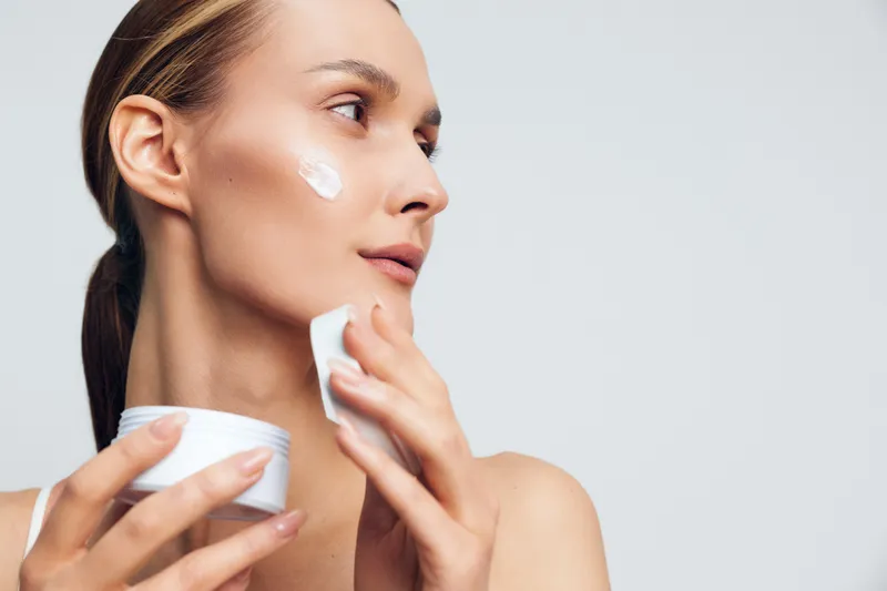 Smiling woman applies facial moisturizer on her cheek, close-up on a soft white background, skincare concept, wellness, and self care mood, soft studio lighting creates warmth
