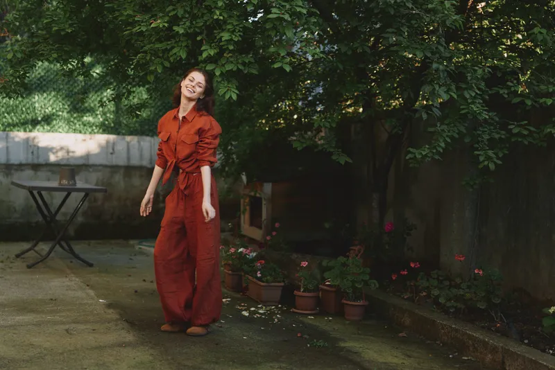 Young woman wearing an orange jumpsuit strolls through a sunlit garden, laughing softly with relaxed posture while greenery, potted flowers, and a rustic wall frame a calm, candid outdoor moment