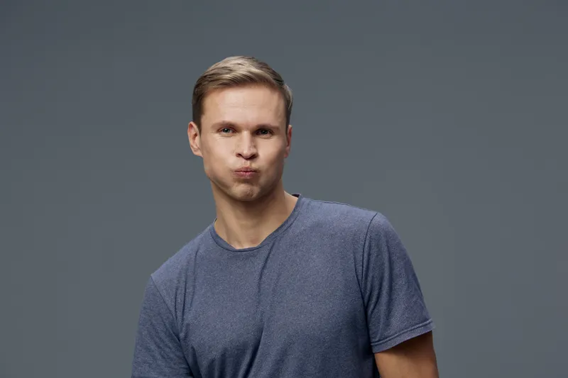 Smiling young man with light brown hair wearing a casual blue t-shirt making a playful kissy face on a plain gray background studio portrait. People lifestyle concept