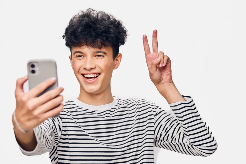 Smiling young man in striped shirt taking selfie on smartphone, showing peace sign, isolated on white background with cheerful expression and curly hair.