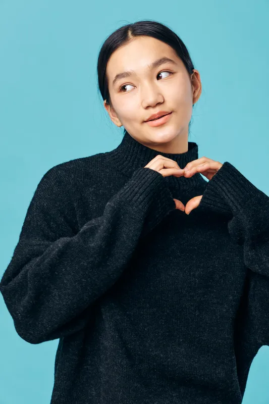 Woman with black sweater making heart shape gesture with hands, looking sideways with gentle smile on blue background for love, care, and positive emotions.