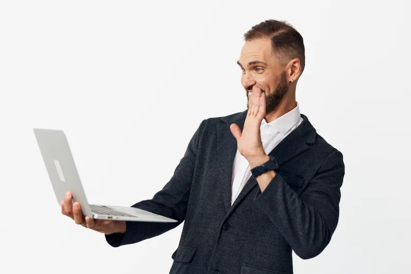 A professional man in a business suit uses a laptop, standing against a bright colored background, conveying focused attention, curiosity, and a friendly, confident smile in a moment of remote work.