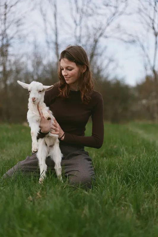A woman kneels on fresh grass, gently holding a curious goat kid in a sunny meadow. A warm lifestyle moment featuring animal companionship and peaceful countryside scenery.