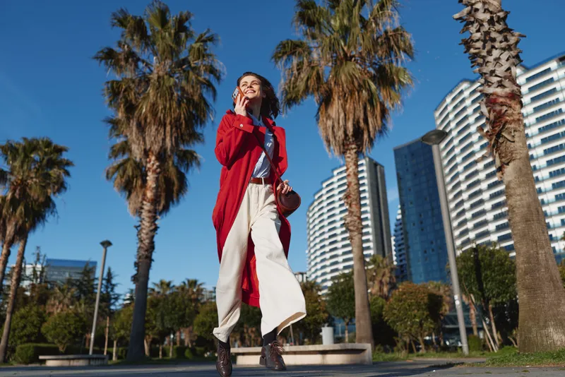 A confident young woman in a red coat walks outdoors on a sunny day with palm trees and modern buildings in the background. Concept of urban lifestyle and fashion.