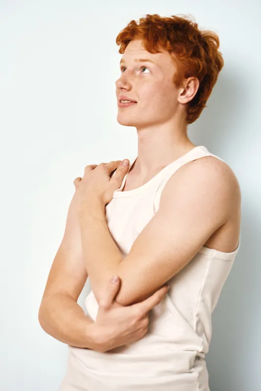 Redhead young man with freckles looking up in white sleeveless shirt smiling and posing with hands on shoulders in bright studio isolated on light background.