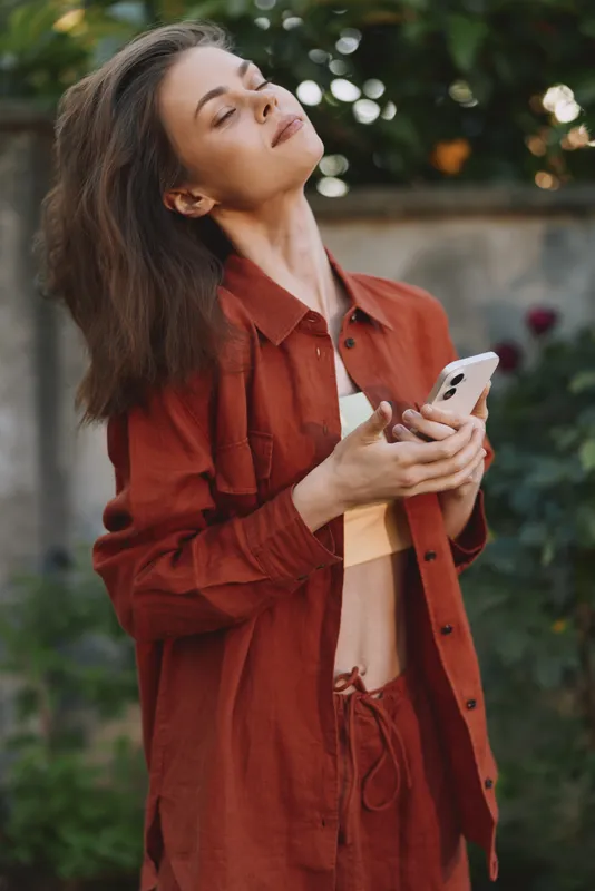 A woman in a rust jacket stands outdoors, looking at a smartphone with a serene expression, capturing a casual lifestyle moment in a sunlit garden