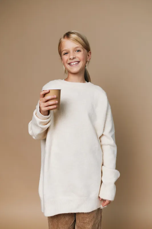 Authentic candid image of a smiling child in a soft white sweater holding a cup, portraying a simple school and lifestyle moment with warm, natural styling.