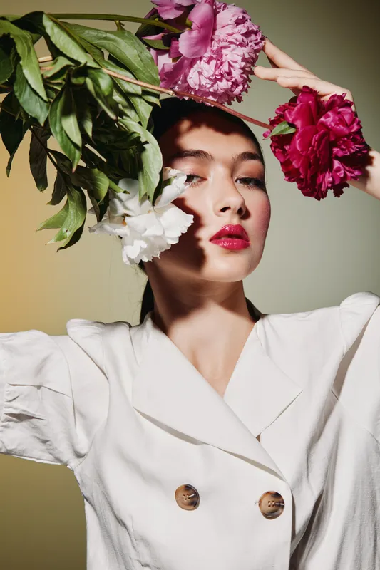 Fashionable woman with glossy red lips poses creatively with vibrant pink flowers against a soft glowing background in a studio. Concept for beauty, fashion, elegance, and floral styling.