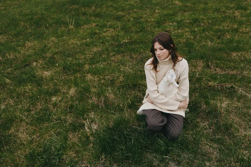 A woman kneels on green grass outdoors wearing a cozy beige sweater, gently holding a white rabbit, capturing a serene lifestyle moment in soft natural light and open field.