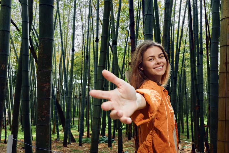 Woman in bamboo forest reaches hand toward camera, smiling in orange jacket. Portrait of young woman connecting with nature, walkway among tall green bamboo stalks.