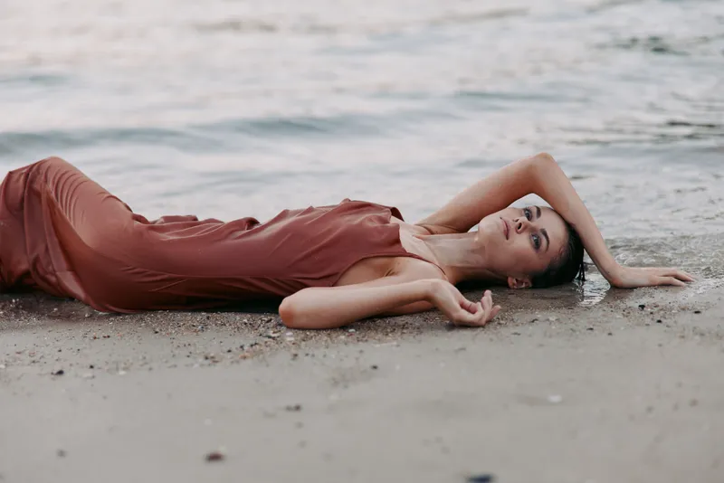 A woman lies on a sandy beach in a flowing dress, posed gracefully with one arm above her head, creating a serene, intimate mood on the shore.