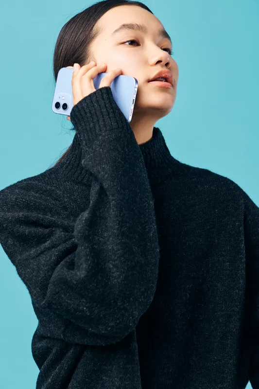 Woman with smartphone talking on mobile device, wearing black sweater and looking aside on blue background. Communication, technology and casual style with modern phone and conversation.