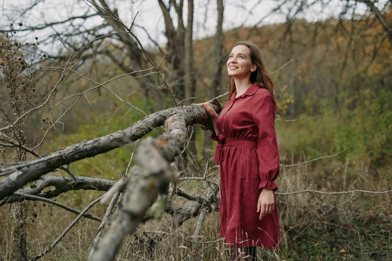 A genuine outdoor portrait of a smiling woman in a red dress among bare trees, capturing authenticity and credible connection with nature in a candid, realistic lifestyle moment.