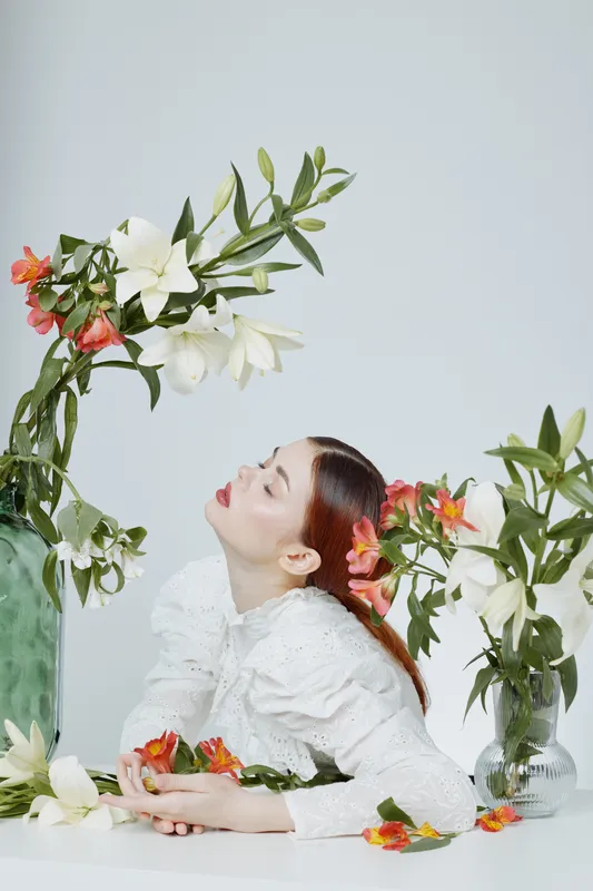 Elegant model in white blouse surrounded by vibrant flowers, showcasing tranquility and beauty in a minimalist floral arrangement