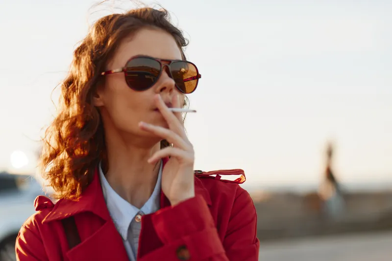 Young woman with curly hair wearing sunglasses and a red coat smokes a cigarette outdoors during golden hour with a blurred urban background.