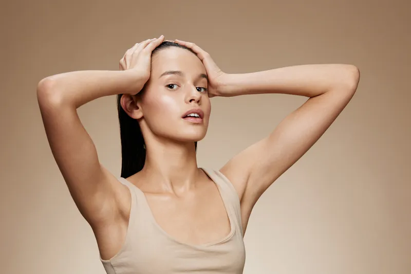 Young woman with long hair expressing tranquility, wearing a neutral tank top against a beige background, symbolizing beauty and wellness