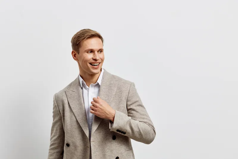 Smiling young man in stylish beige blazer and white shirt showing confident expression on plain white background studio portrait. People lifestyle concept
