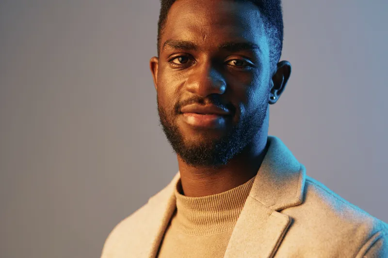 Stylish young man with a confident smile, wearing a beige blazer and turtleneck, set against a soft blue and orange gradient background