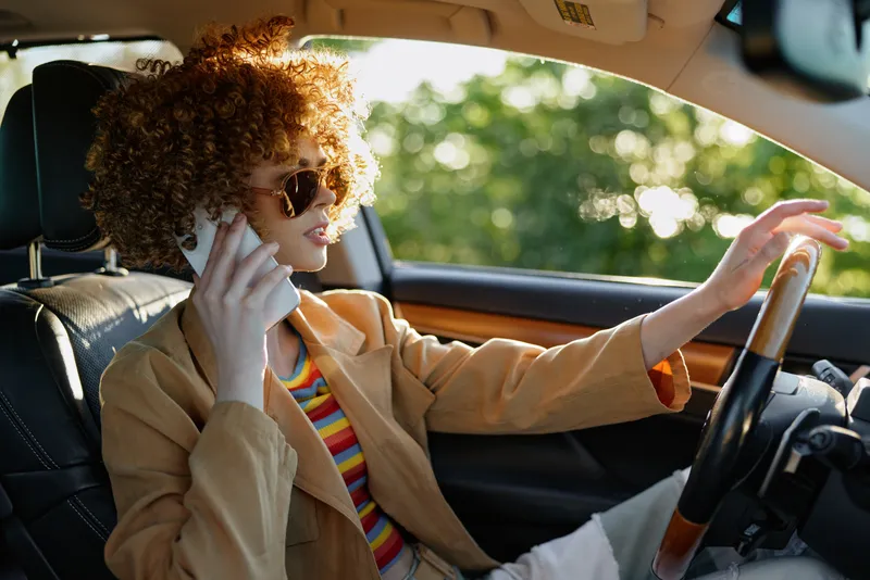 Vibrant woman driving a car, wearing sunglasses and a beige blazer, curly hair, striped top, retro film color tones, capturing a carefree lifestyle moment on the sunny road