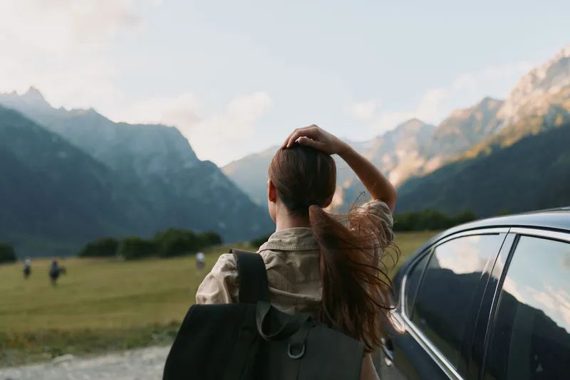 A young woman with a backpack stands beside a car, gazing at distant mountains, embodying travel, discovery and anticipation.