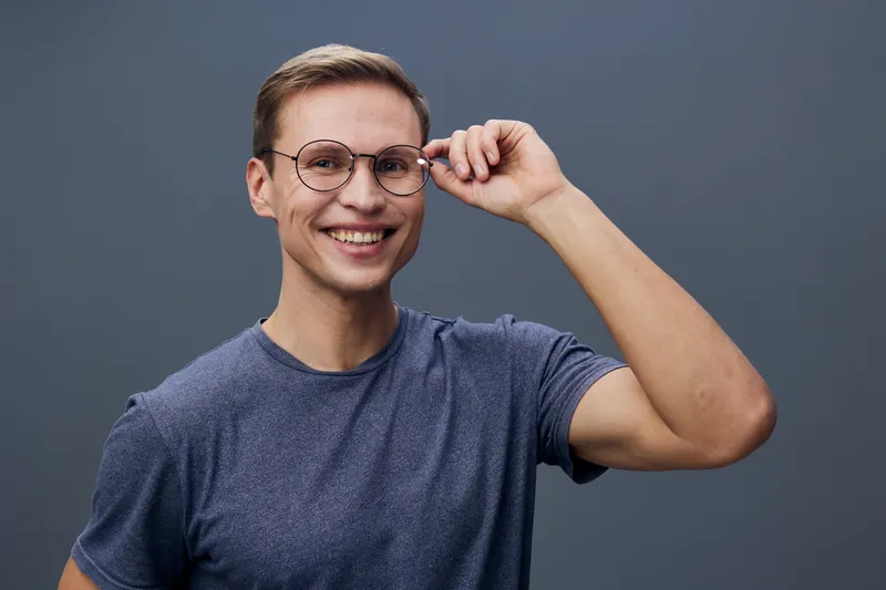 Confident young man wearing glasses and casual grey t-shirt smiles at camera on plain grey background studio portrait. People lifestyle concept
