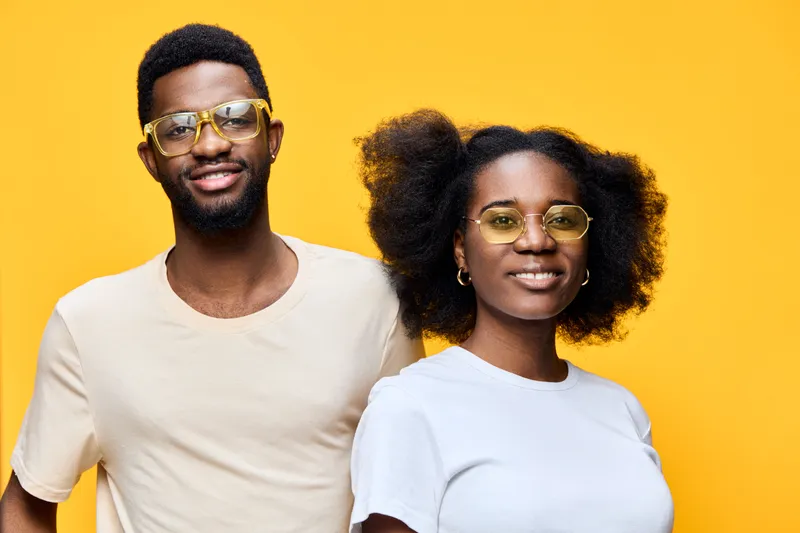 Young African couple smiling together against a vibrant yellow background, wearing casual outfits and glasses, capturing joy and confidence
