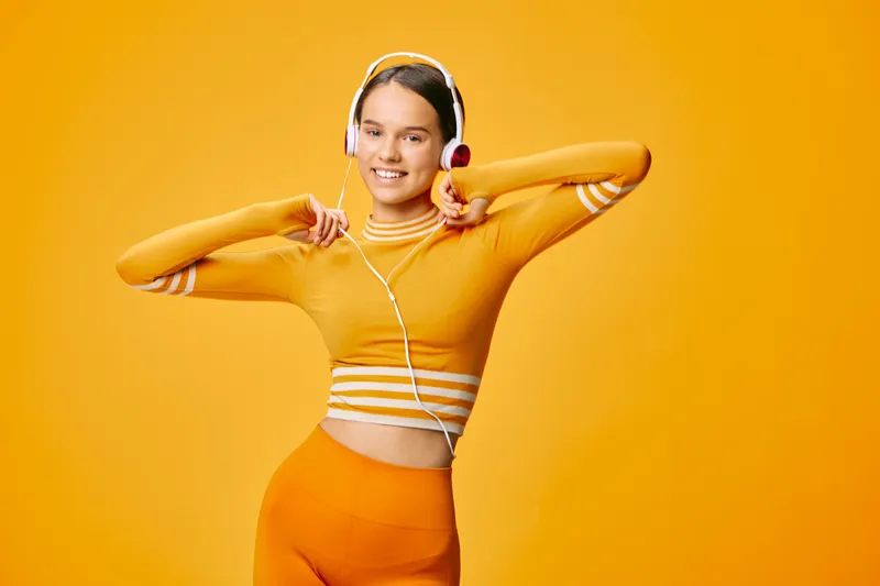 Fitness and sport themed portrait of a smiling woman in orange athletic wear with headphones, posing energetically in a bright studio, showcasing movement and motivation for workout and healthy