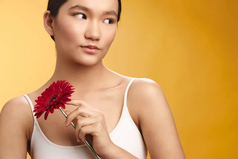 Asian woman holds a red flower against a warm backdrop, gazing softly aside. The simple scene conveys calm beauty and graceful poise, inviting a gentle, intimate mood perfect for lifestyle and