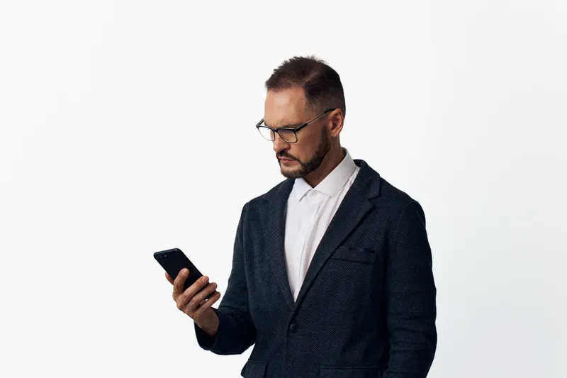 A professional man in a dark patterned suit stands against a clean white backdrop, focused on his smartphone, conveying confidence and contemplation.