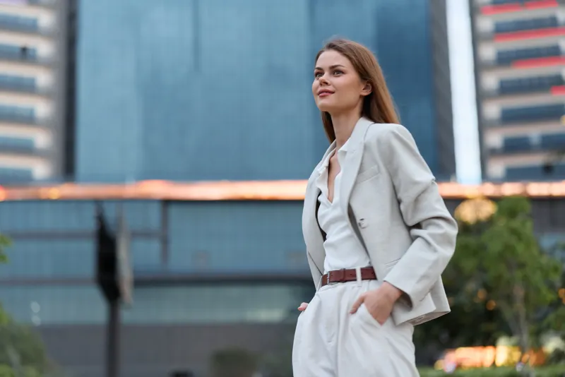 confident woman in white business suit walking outdoors in urban environment with modern glass buildings and natural evening light, showing empowerment and professionalism