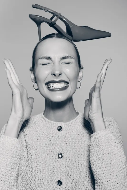 Young white woman with fair skin smiling with closed eyes holds high heel shoe on head in studio. Concept of fashion and playful mood.