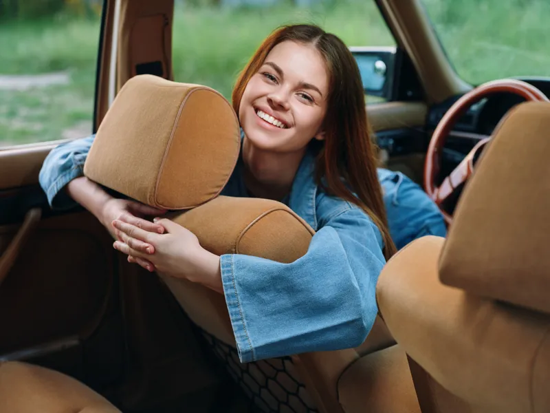 Happy young woman with long hair smiling inside a vintage car, wearing a denim jacket, conveying joy and summer vibes in a natural outdoor setting.