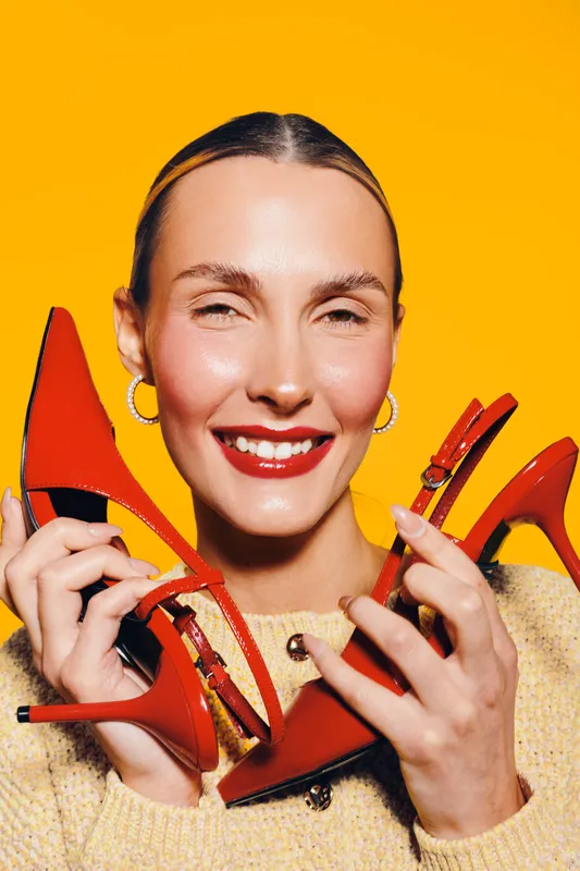 Smiling woman with red lipstick holds red high heel shoes on bright yellow background. Concept of fashion, style, accessories, and joyful expression in studio lighting.